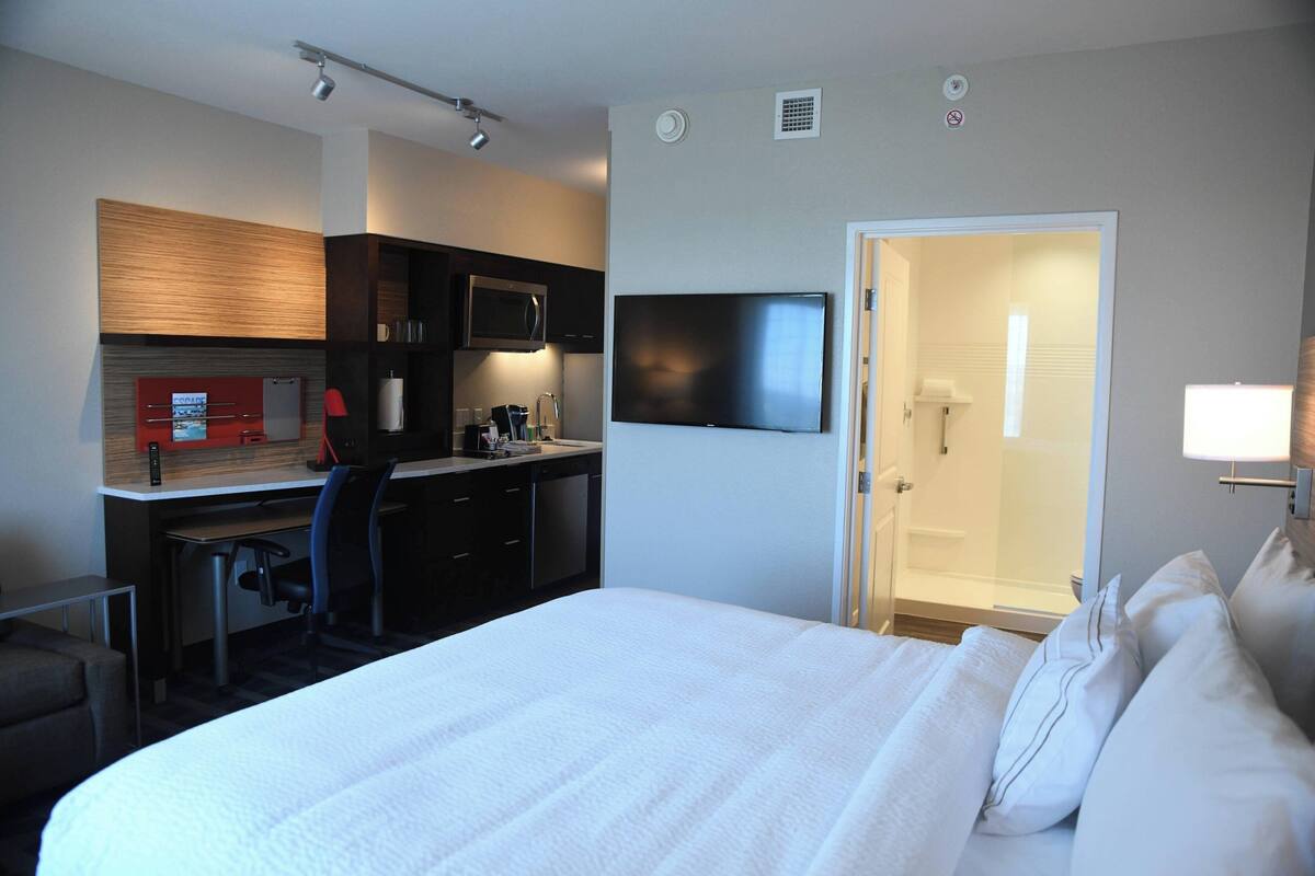 A hotel room with a bed, a desk with a chair, a microwave, a TV, and a door leading to a bathroom.