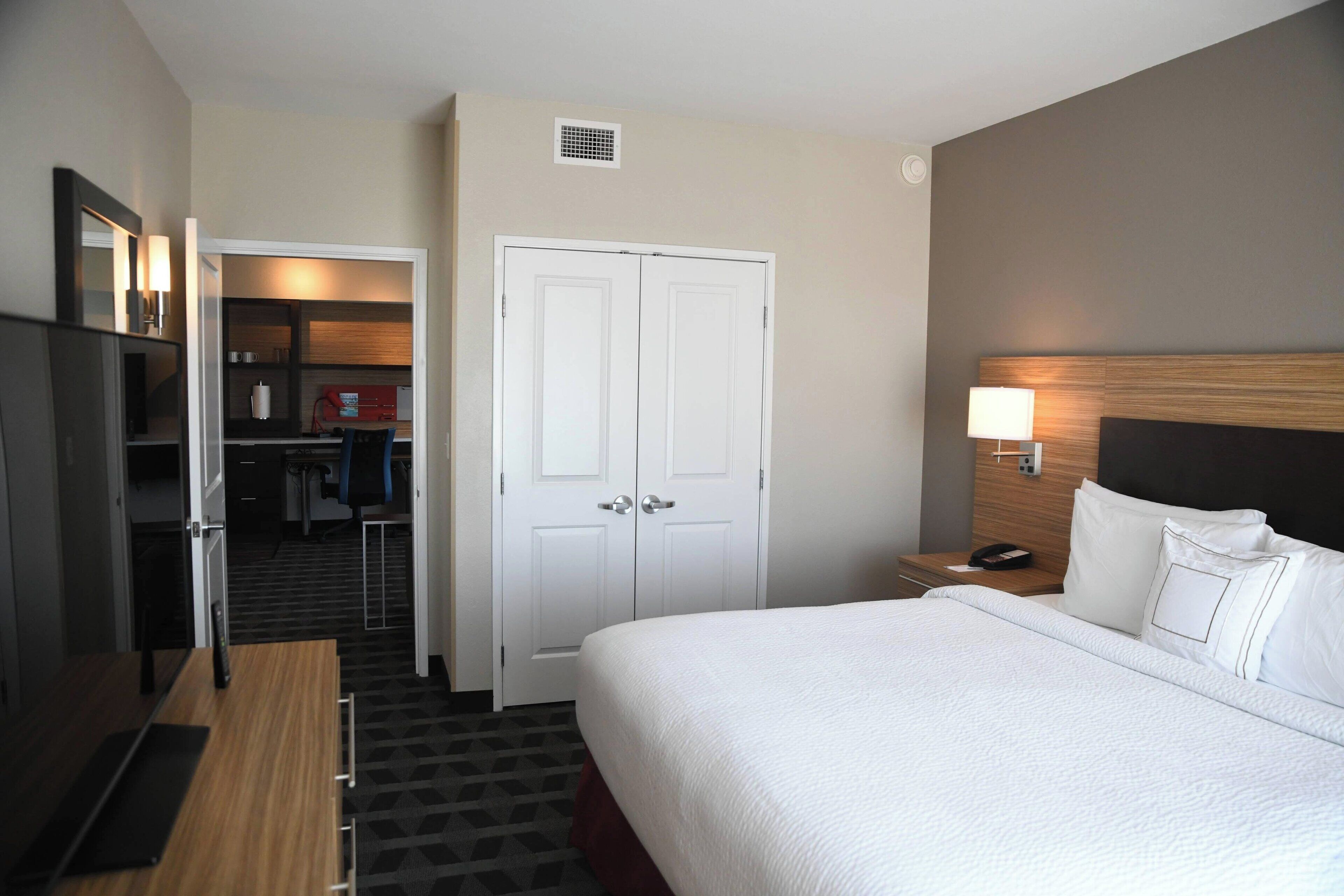A hotel room with a bed, a desk, a chair, a wardrobe, and a nightstand with a lamp.