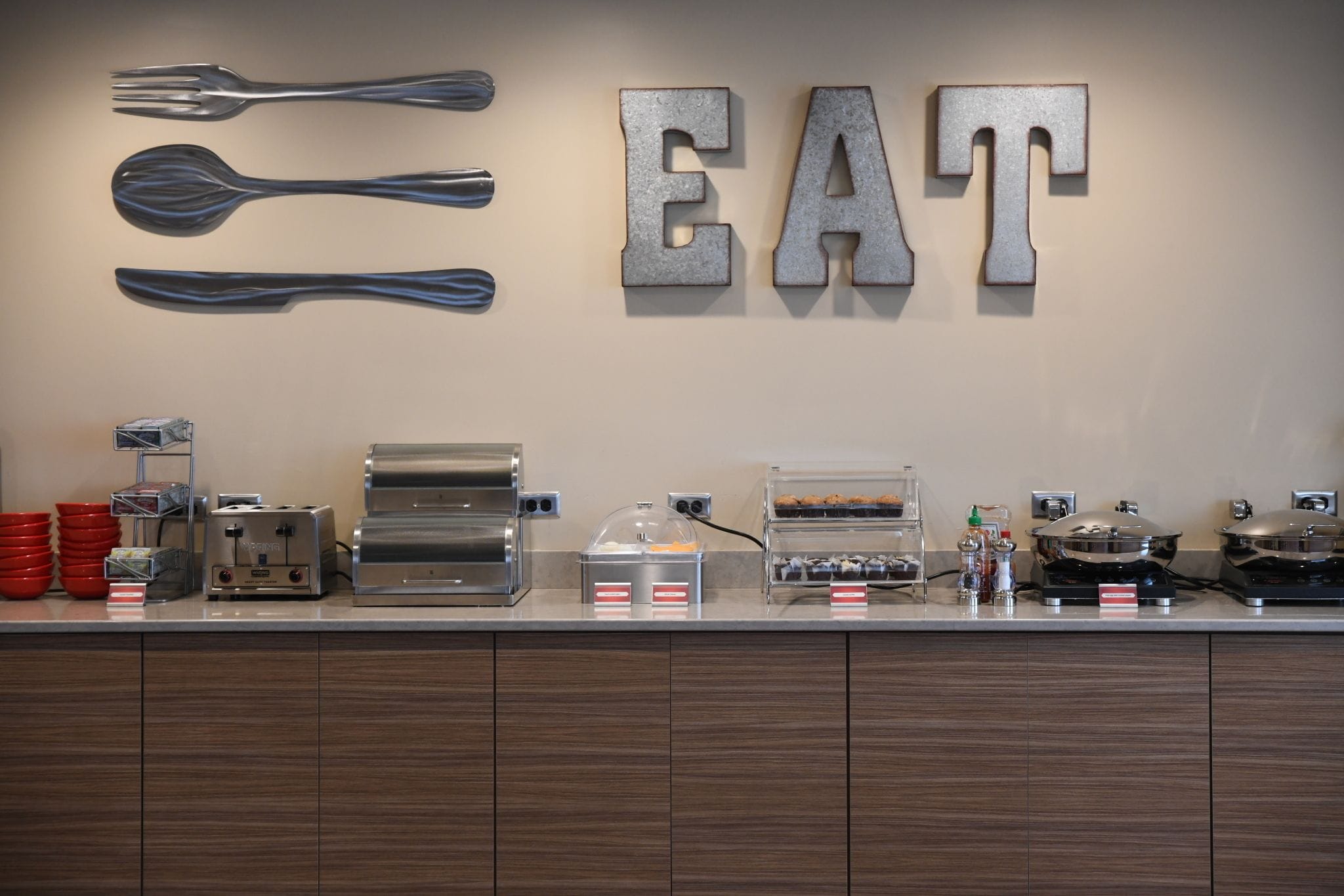 A buffet counter with utensils and the word "EAT" on the wall, a toaster, a coffee machine, and a variety of food items.