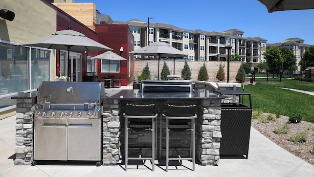 Un espace bar extérieur doté d’un barbecue en acier inoxydable, de deux tabourets de bar et de parasols.