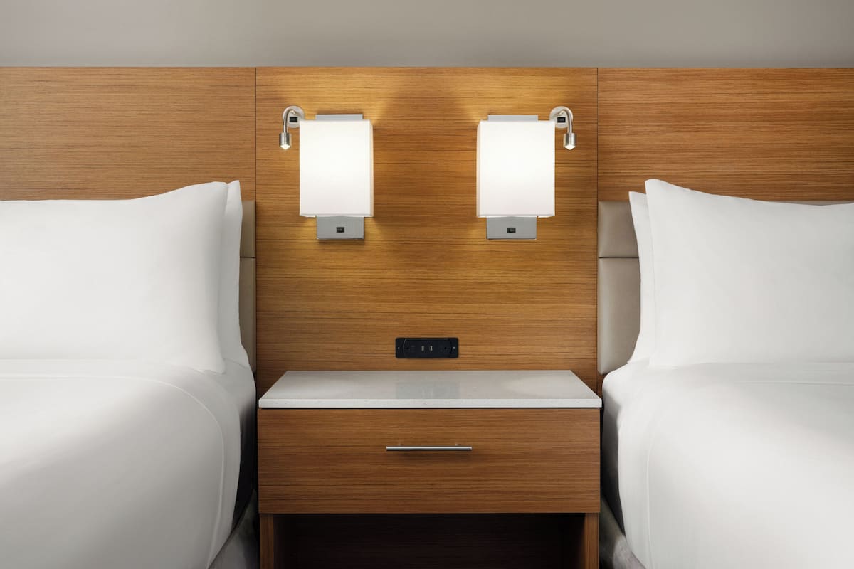 A hotel room with two beds, a wooden headboard, a nightstand with a drawer, and two wall-mounted lamps.