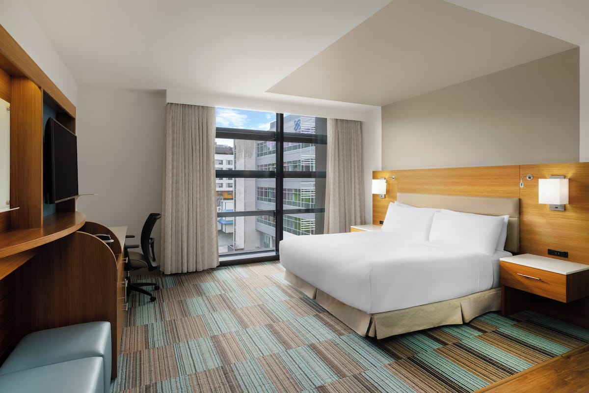 A hotel room with a large bed, a desk with a chair, a television, and a view of the city through a large window.