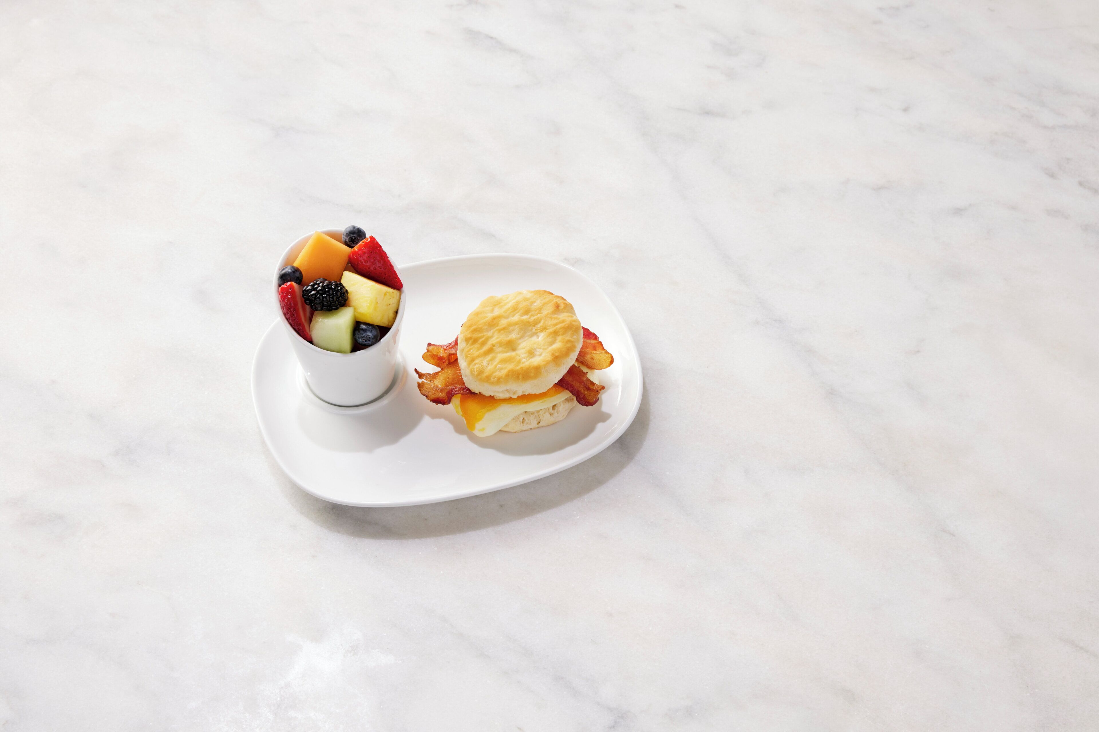 A white plate with a fruit bowl and a breakfast sandwich on a marble surface.