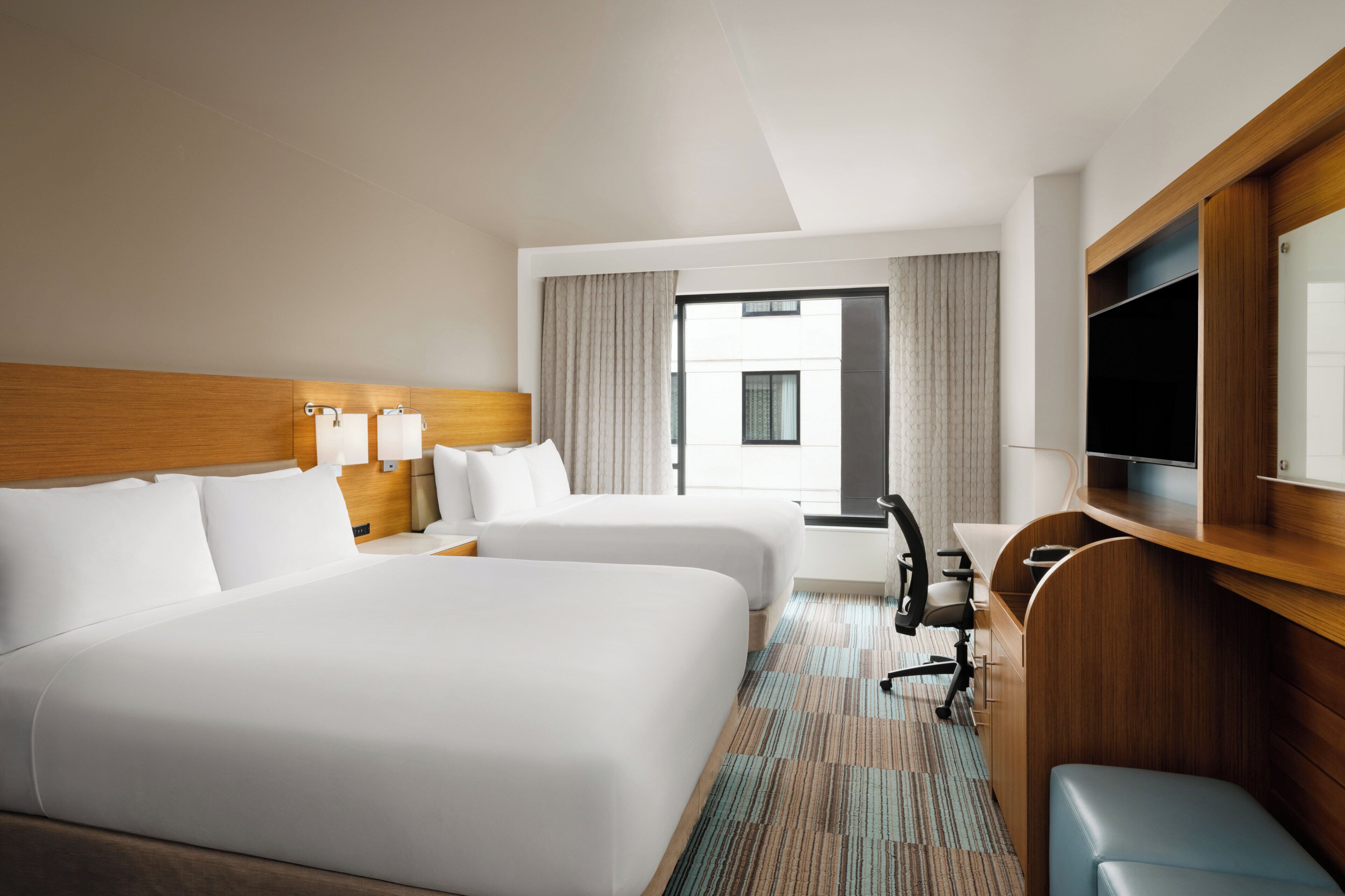 Une chambre d’hôtel avec deux lits, un bureau, une chaise, une télévision et une grande fenêtre.