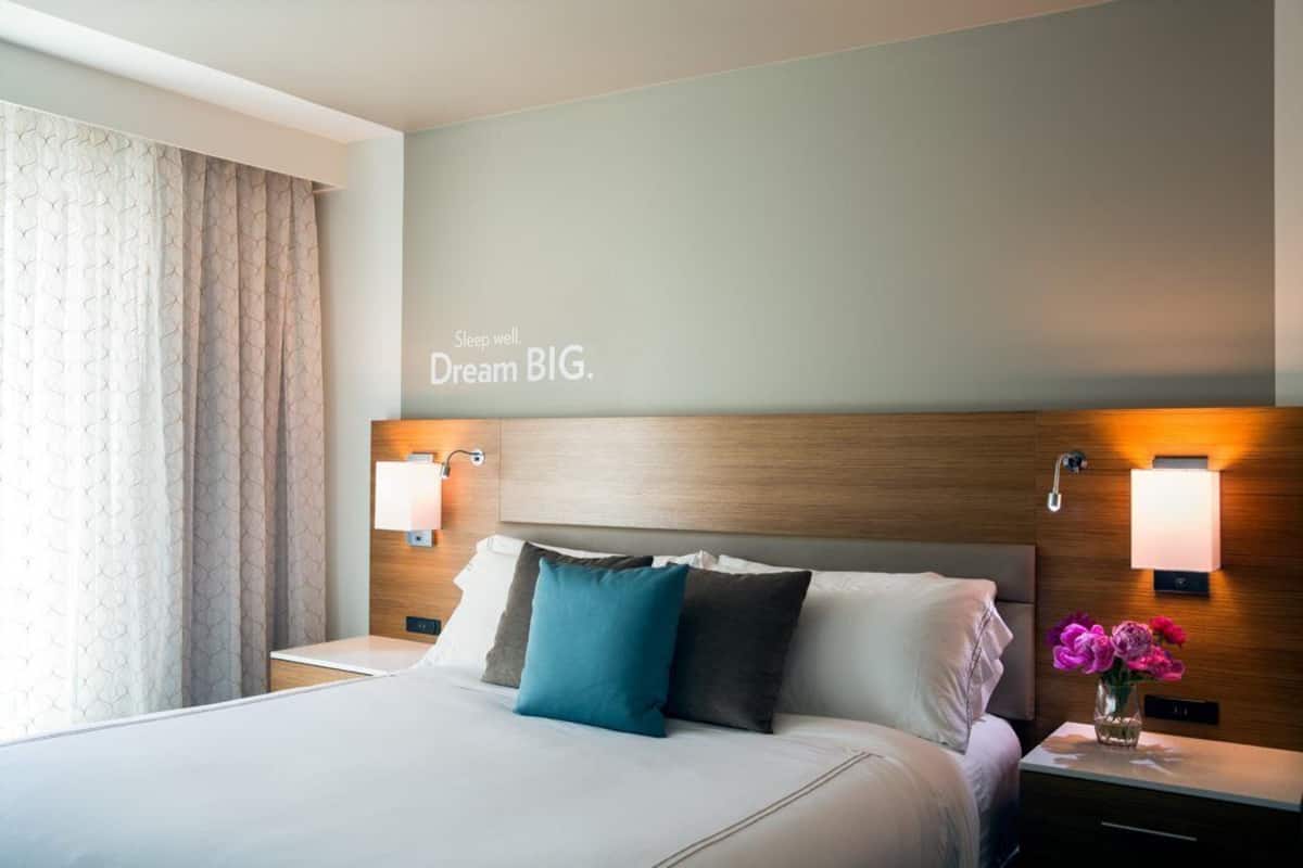 A hotel room with a large bed, two bedside tables, a nightstand, a wall-mounted light, and a decorative wall with a motivational quote.