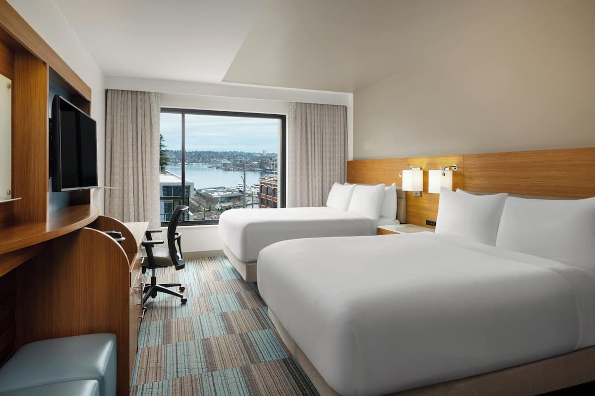 A hotel room with two beds, a desk, a chair, a TV, and a large window with a city view.
