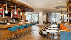 A modern bar area with a curved wooden counter, blue bar stools, and round tables with orange cushions.