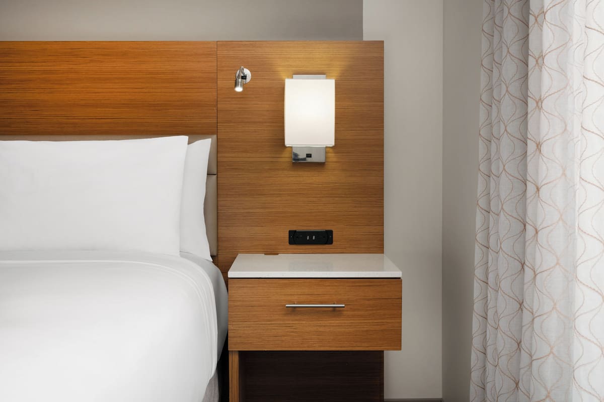 A hotel room with a wooden headboard, a bedside table with a lamp, a drawer, and a patterned curtain.