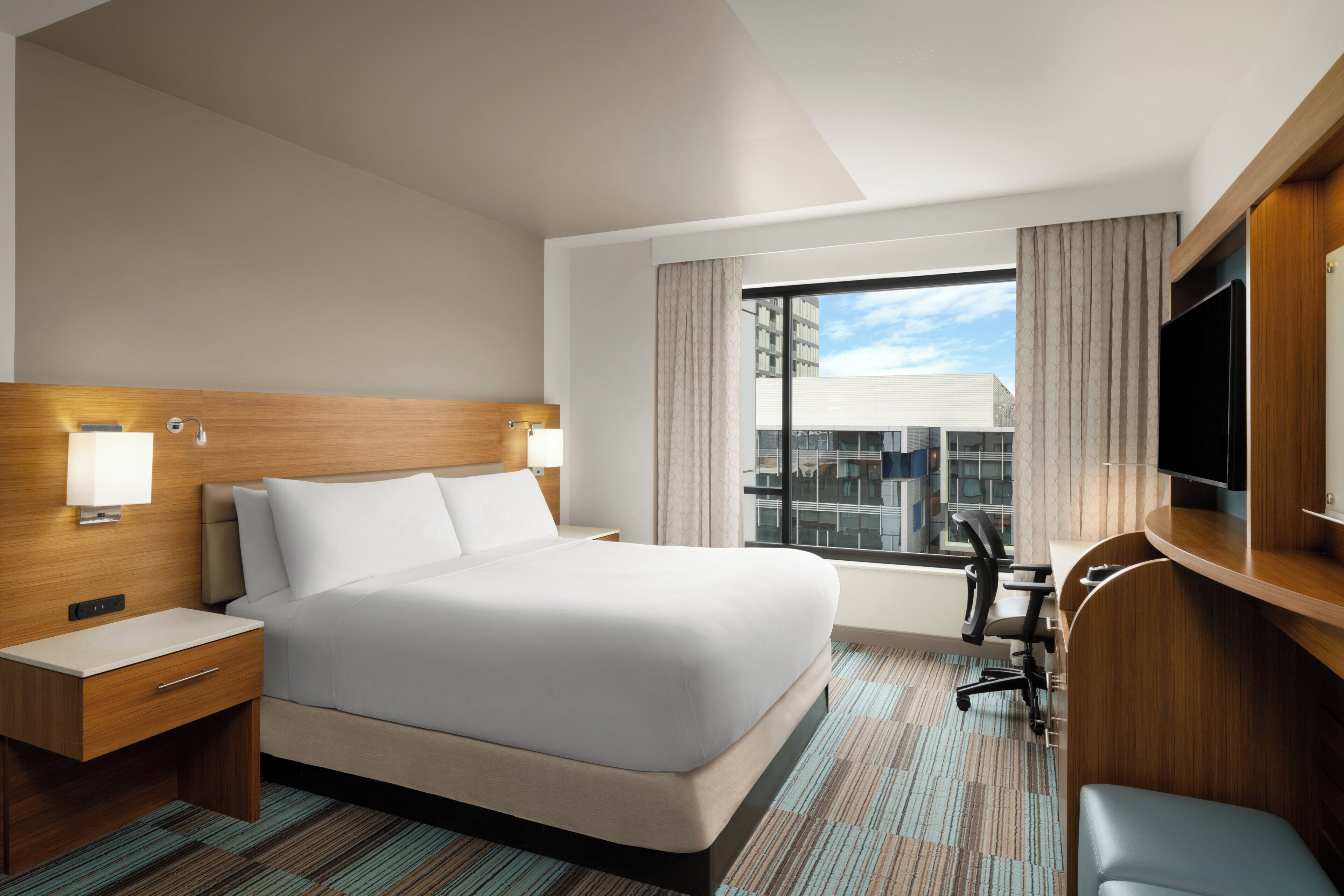 A hotel room with a large bed, a desk, a chair, a television, and a window with curtains.