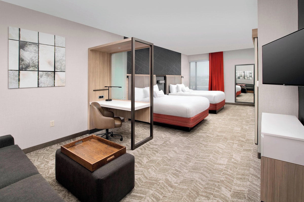 A modern hotel room with a desk, two beds, a TV, and a large window.