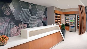 Uma área de recepção com design moderno, apresentando um padrão geométrico na parede, um balcão de madeira, uma planta em vaso e uma geladeira com bebidas.