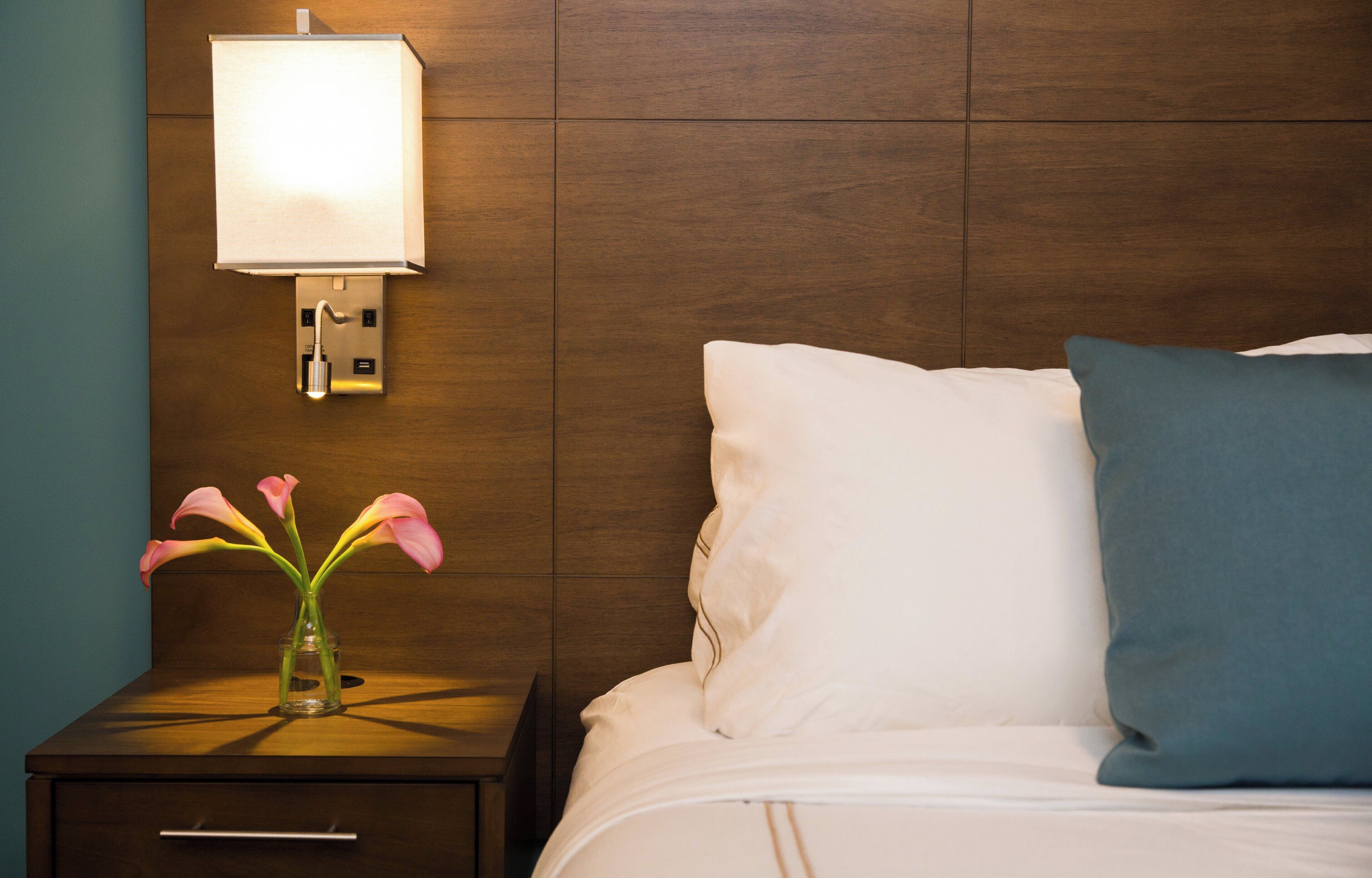 A hotel room with a wooden headboard, a bedside table with a lamp, a vase with flowers, and a bed with pillows.