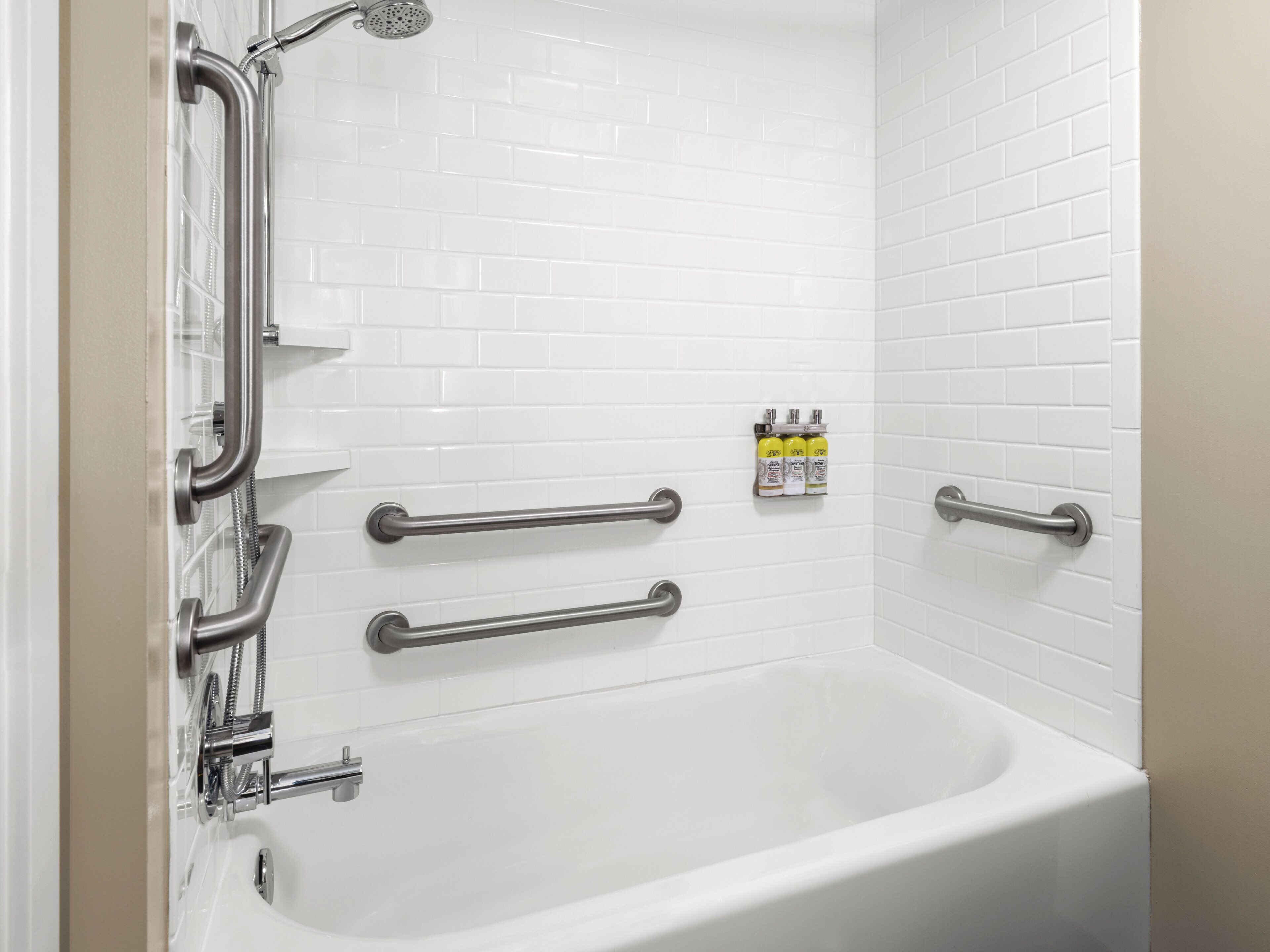 Un baño con azulejos blancos, una bañera, barras de apoyo y un cabezal de ducha.