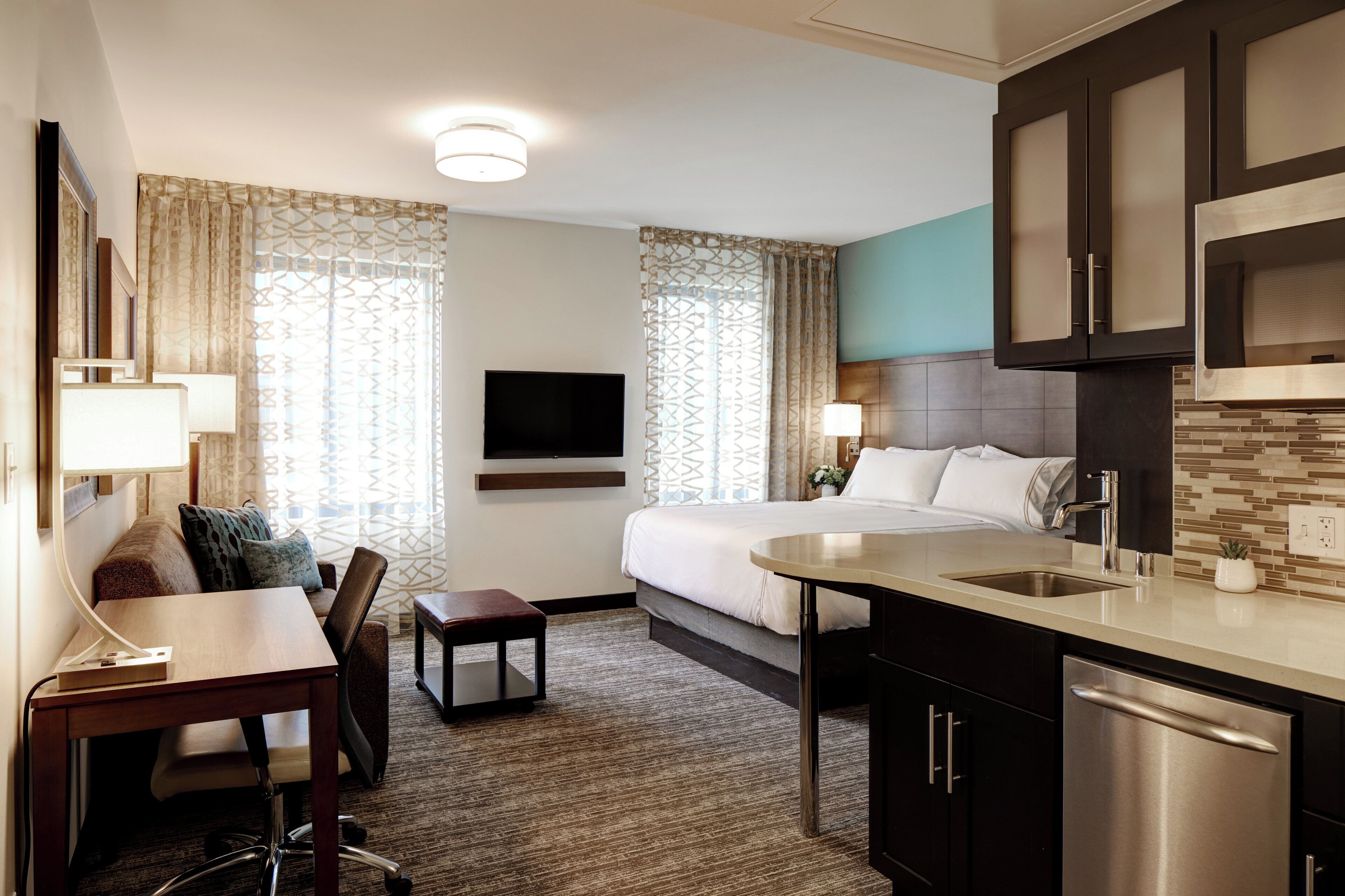 A hotel room with a bed, a desk, a chair, a TV, a kitchen area with a sink and a dishwasher, and patterned curtains.