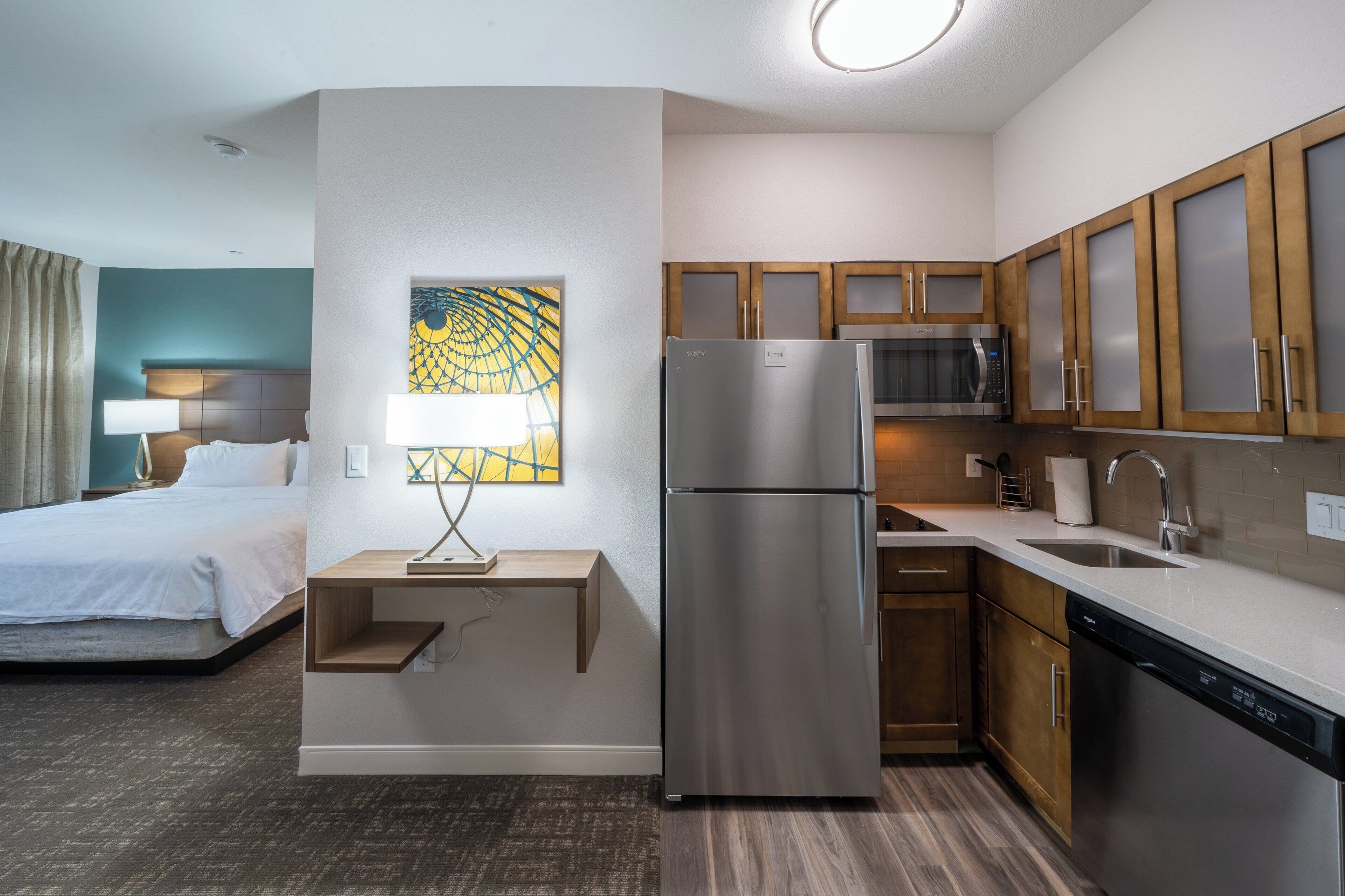 A compact hotel room with a kitchenette, a bed, a desk, and a painting on the wall.