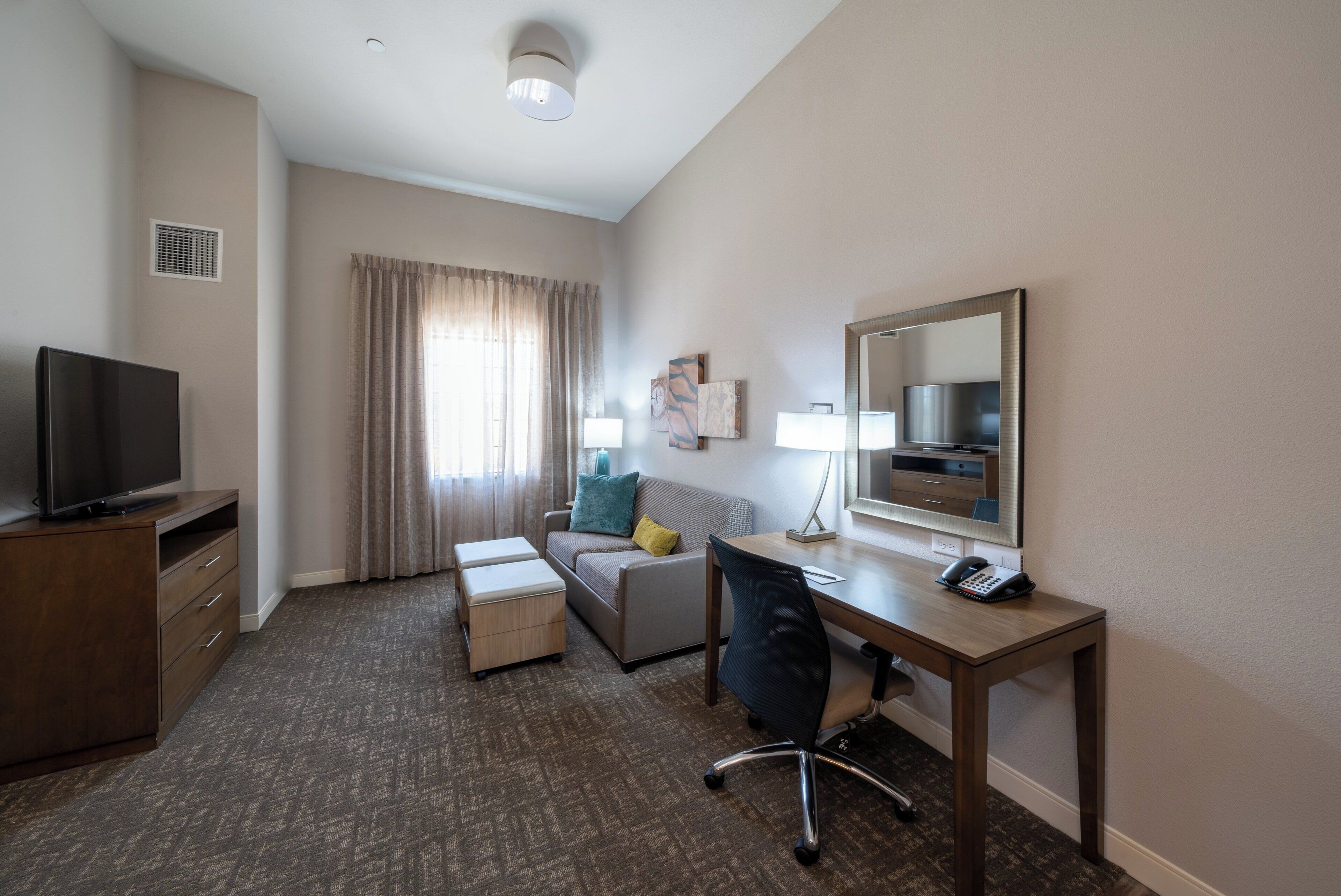 A hotel room with a flat-screen TV, a desk with a telephone, a chair, a sofa, and a window with curtains.