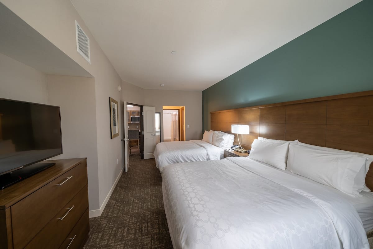A hotel room with a large bed, a TV, a wooden dresser, and a hallway leading to a bathroom.