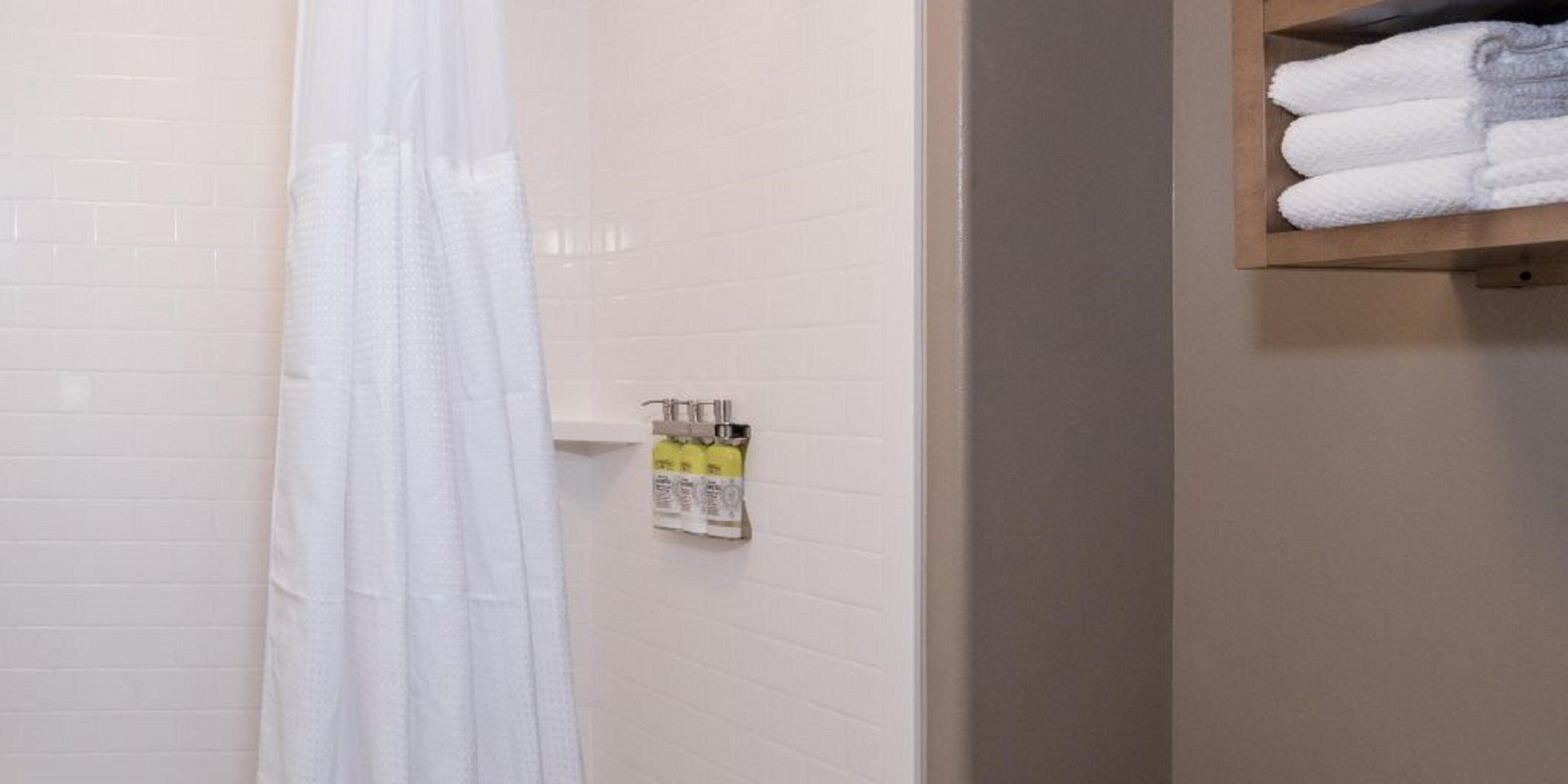 A bathroom with a white tiled wall, a shower curtain, a towel rack with folded towels, and a soap dispenser.