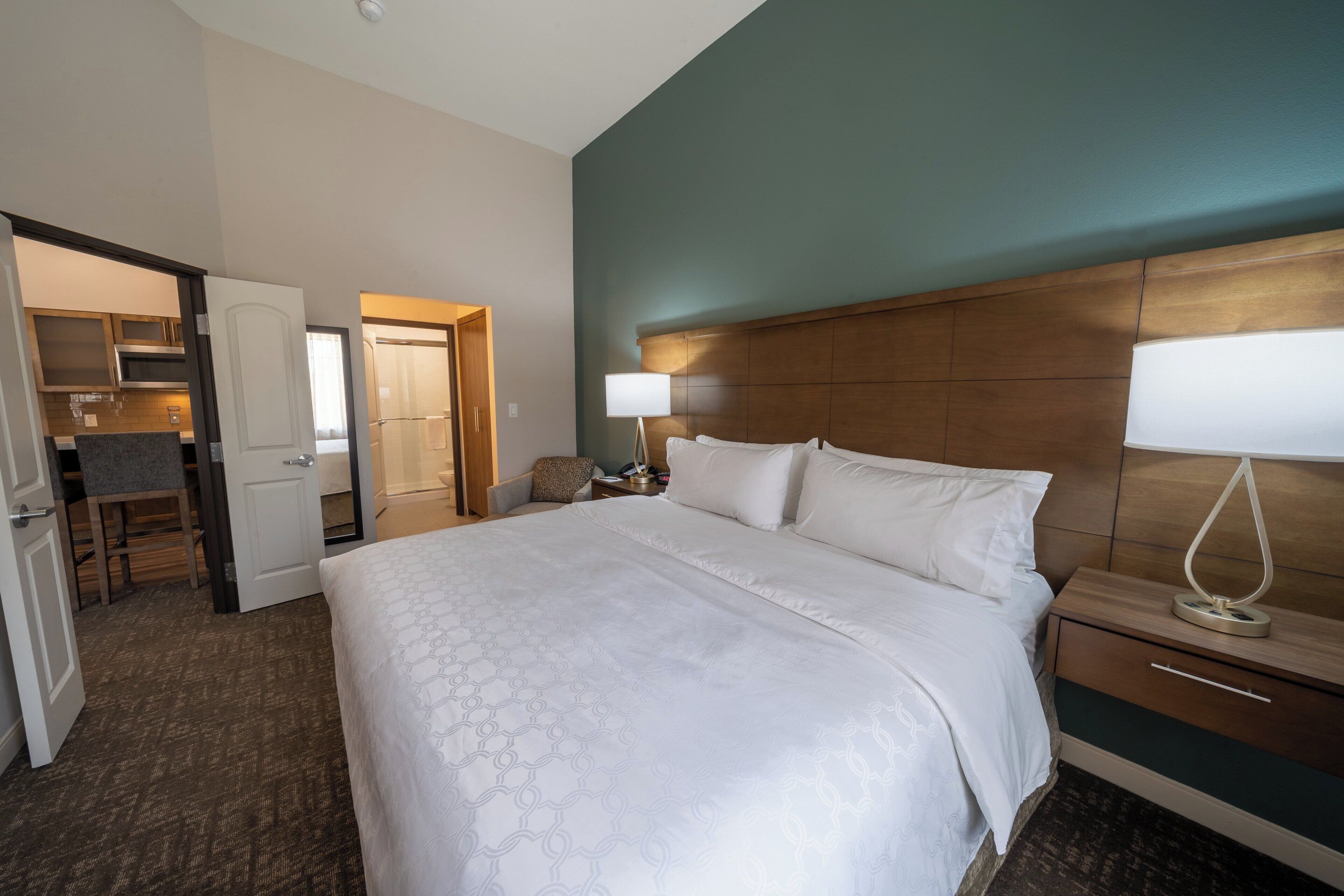 A hotel room with a large bed, a bedside table with a lamp, a wooden headboard, and a view of the bathroom and kitchen area.