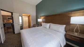 A hotel room with a large bed, a bedside table with a lamp, a wooden headboard, and a view of the bathroom and kitchen area.
