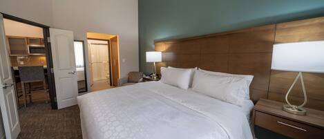 A hotel room with a large bed, a bedside table with a lamp, a wooden headboard, and a view of the bathroom and kitchen area.