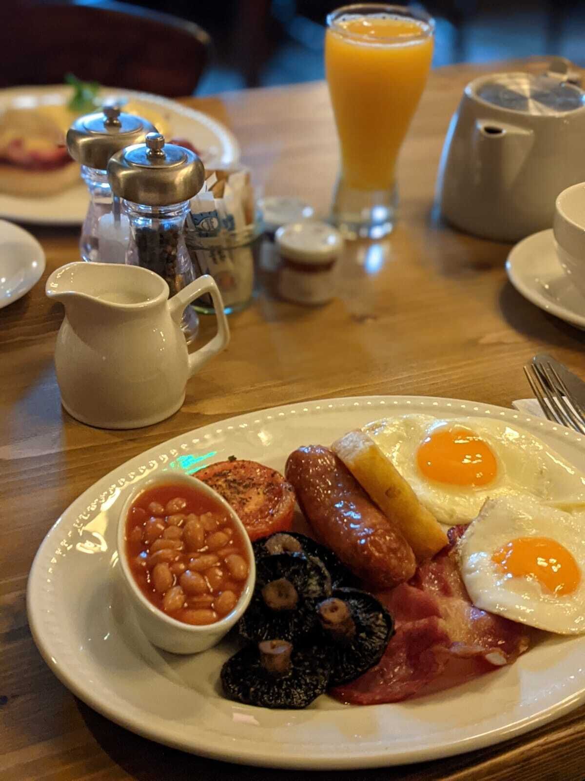 Une assiette de petit-déjeuner comprenant des œufs, des saucisses, des haricots et du boudin noir, accompagnée d’un verre de jus d’orange et d’une carafe de lait, le tout posé sur une table en bois.