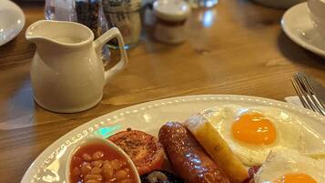 Une assiette de petit-déjeuner comprenant des œufs, des saucisses, des haricots et du boudin noir, accompagnée d’un verre de jus d’orange et d’une carafe de lait, le tout posé sur une table en bois.