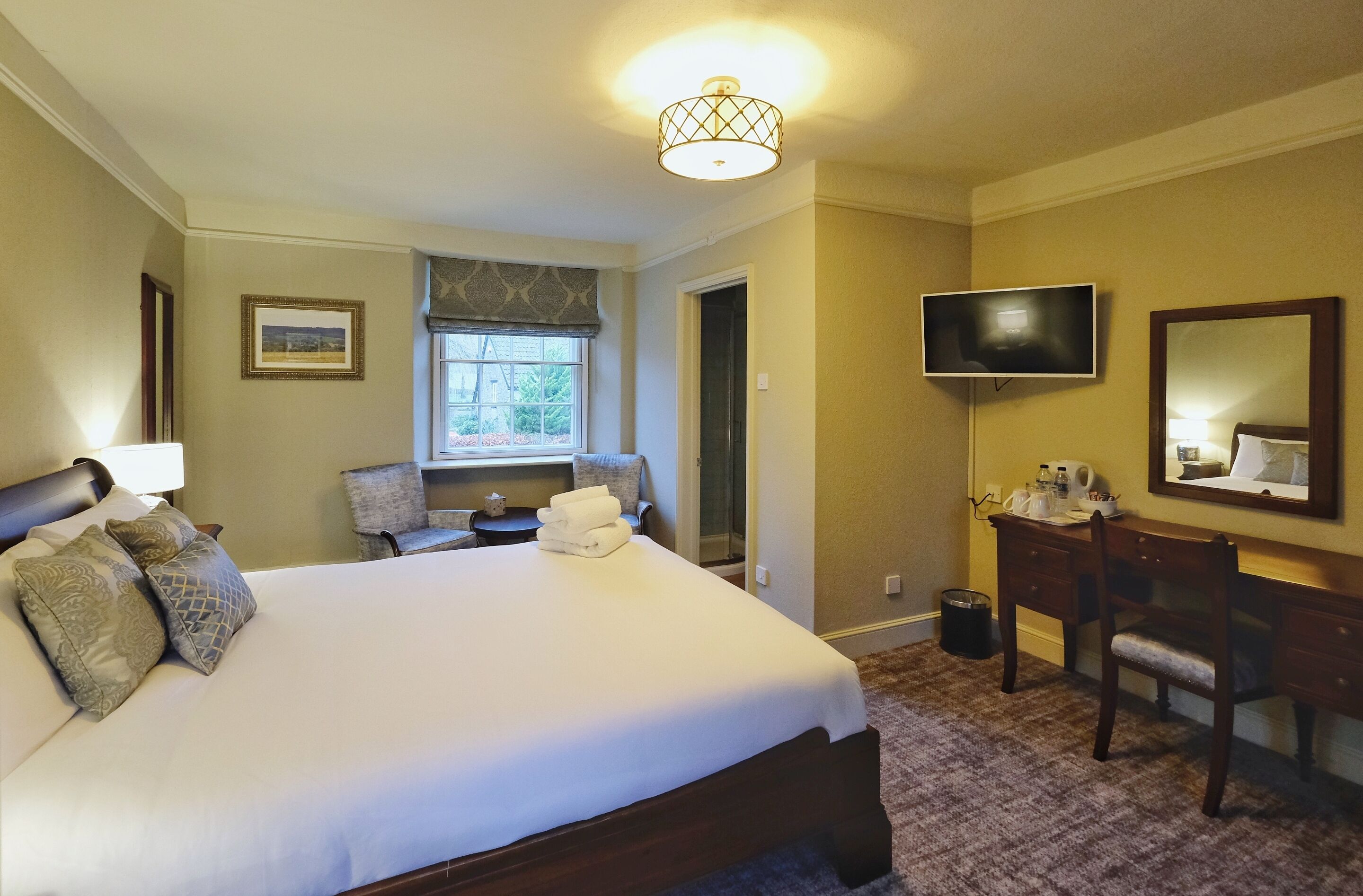 A hotel room with a large bed, a desk with a chair, a television, and a window with a view of trees.