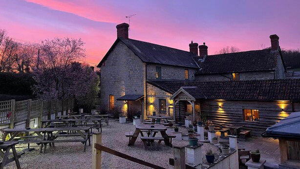 A rustic stone building with a wooden extension, outdoor seating, and a gravel ground. - The Greyhound Inn Taunton (Taunton)