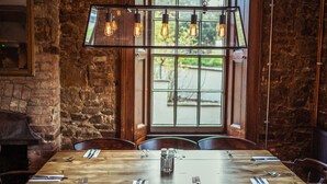 A rustic dining area with a wooden table set for six, a hanging light fixture with exposed bulbs, and a large window with a view of greenery. - The Greyhound Inn Taunton (Taunton)