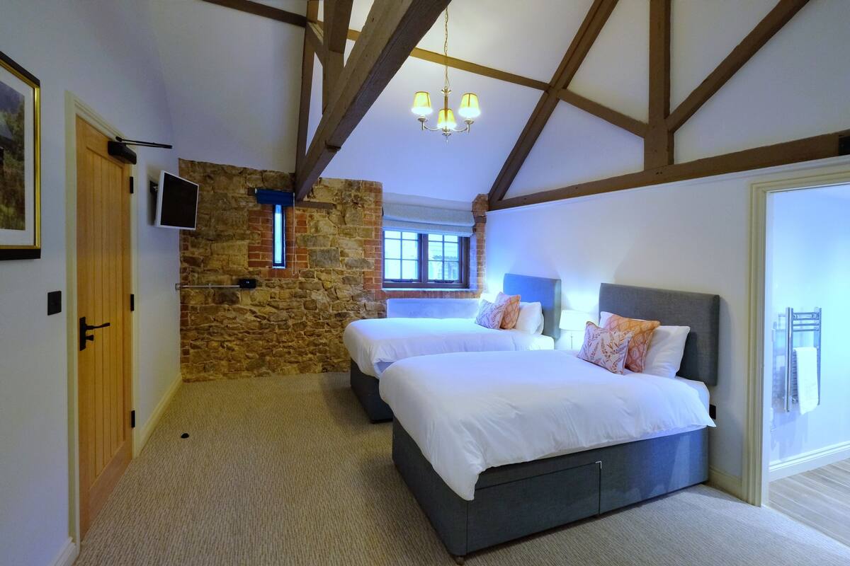 A bedroom with two beds, a stone wall, wooden beams, a chandelier, and a window.