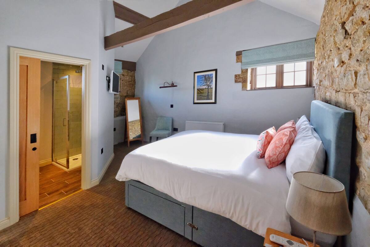 A bedroom with a large bed, a bedside lamp, a stone wall, a framed picture, and a wooden door leading to a bathroom.