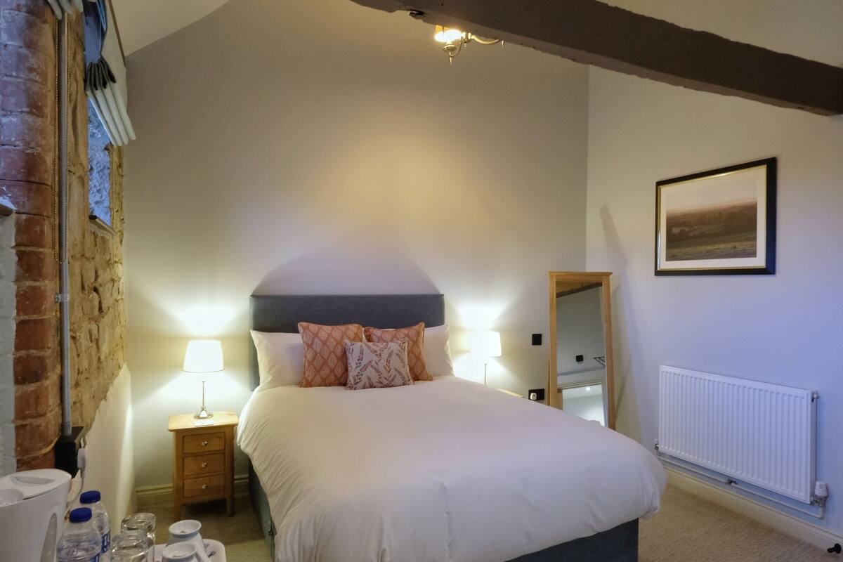 A bedroom with a large bed, wooden beams, a nightstand with a lamp, a radiator, and a framed picture on the wall.