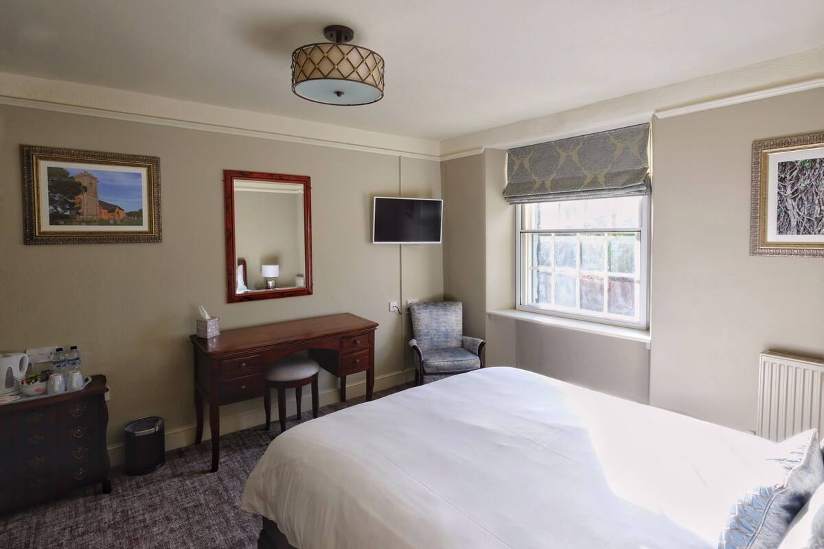 A hotel room with a bed, a desk with a chair, a television, a window with a view, and a radiator.