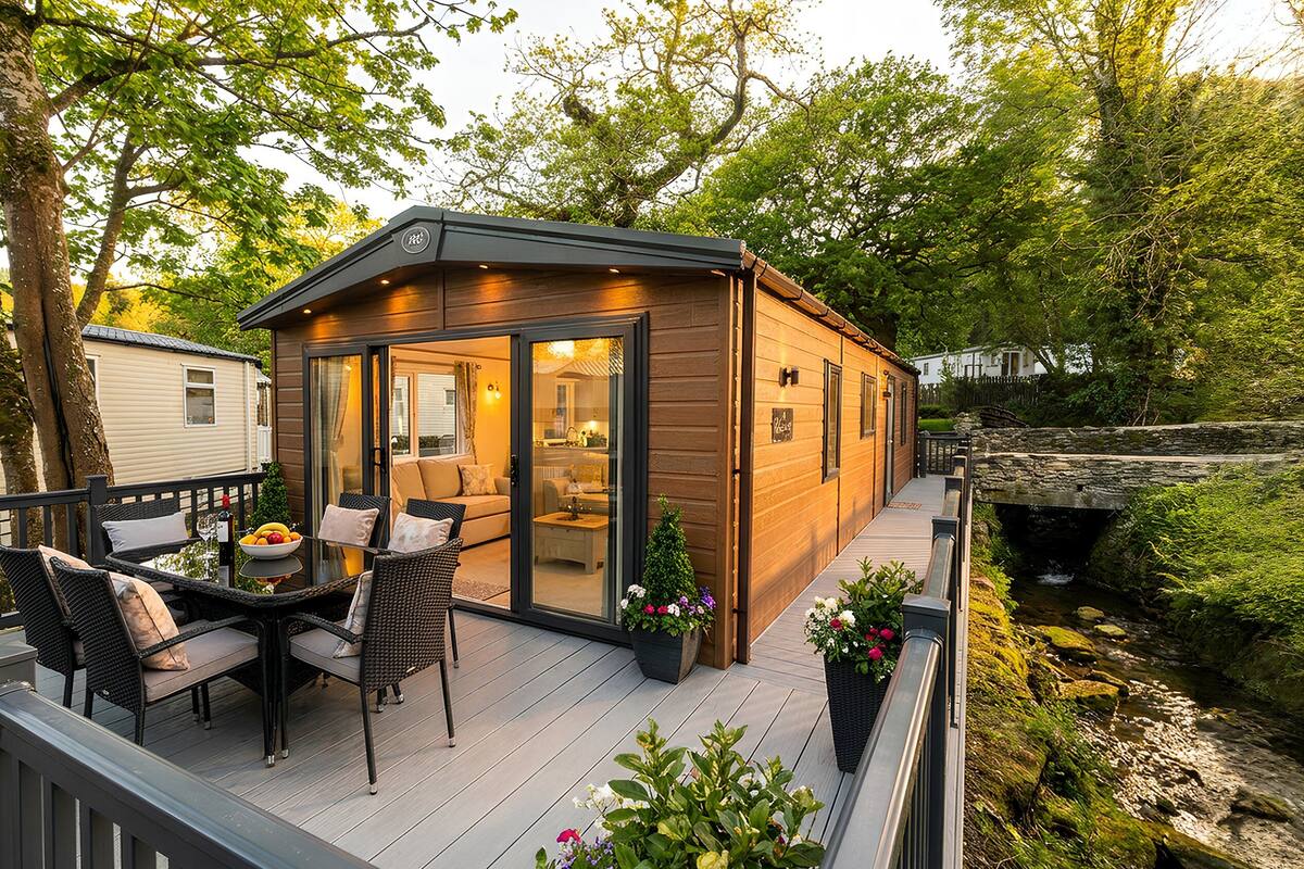 A wooden cabin with a deck, furnished with outdoor seating and a table, overlooking a stream.