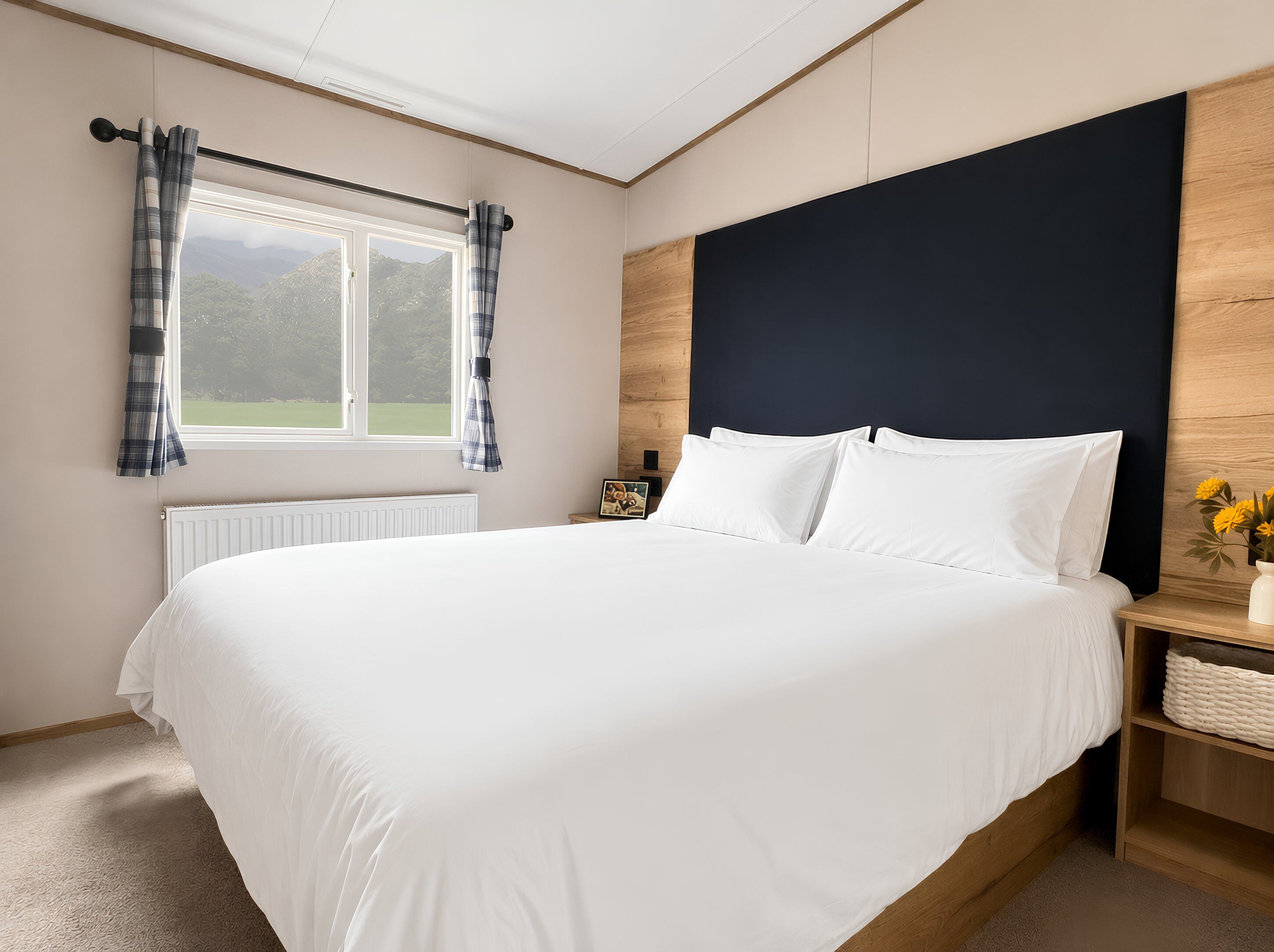 Ein Schlafzimmer mit einem großen Bett, einem Fenster mit Vorhängen, einem Nachttisch mit Lampe und Blick ins Grüne.