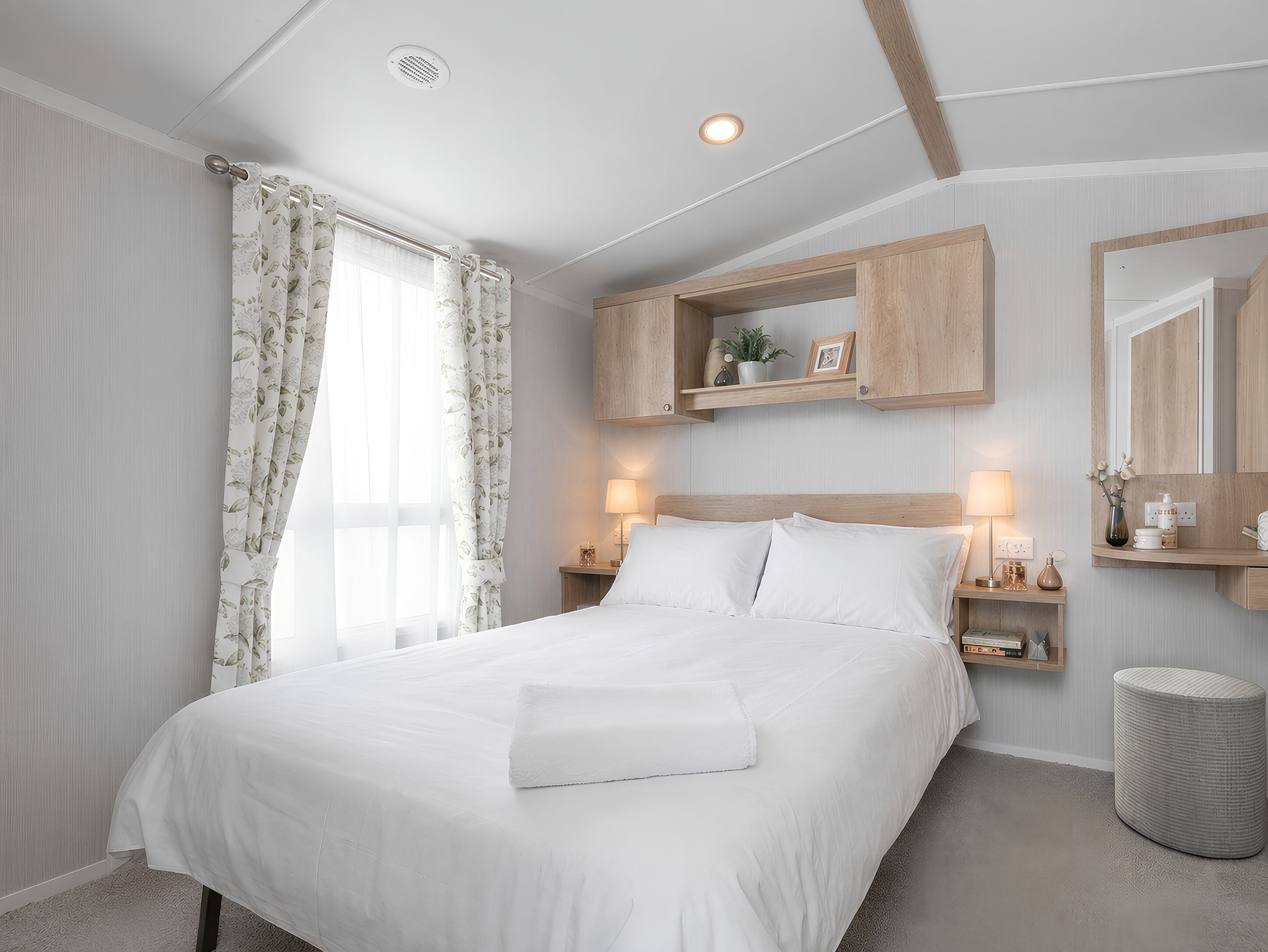 Un lit bien fait, avec du linge de lit blanc, une table de chevet avec une lampe, une petite étagère avec des objets décoratifs et une fenêtre ornée de rideaux à motifs floraux.