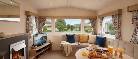 Ein gemütliches Zimmer mit Kamin, Fernseher, einer Couch mit Kissen, einem Esstisch mit Frühstücksgebäck und großen Fenstern mit Vorhängen.