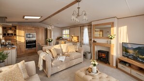 A cozy living room with a sofa, a fireplace, a television, and a kitchen area with wooden cabinets and a stove. - Aberdunant Caravans Direct (Porthmadog)