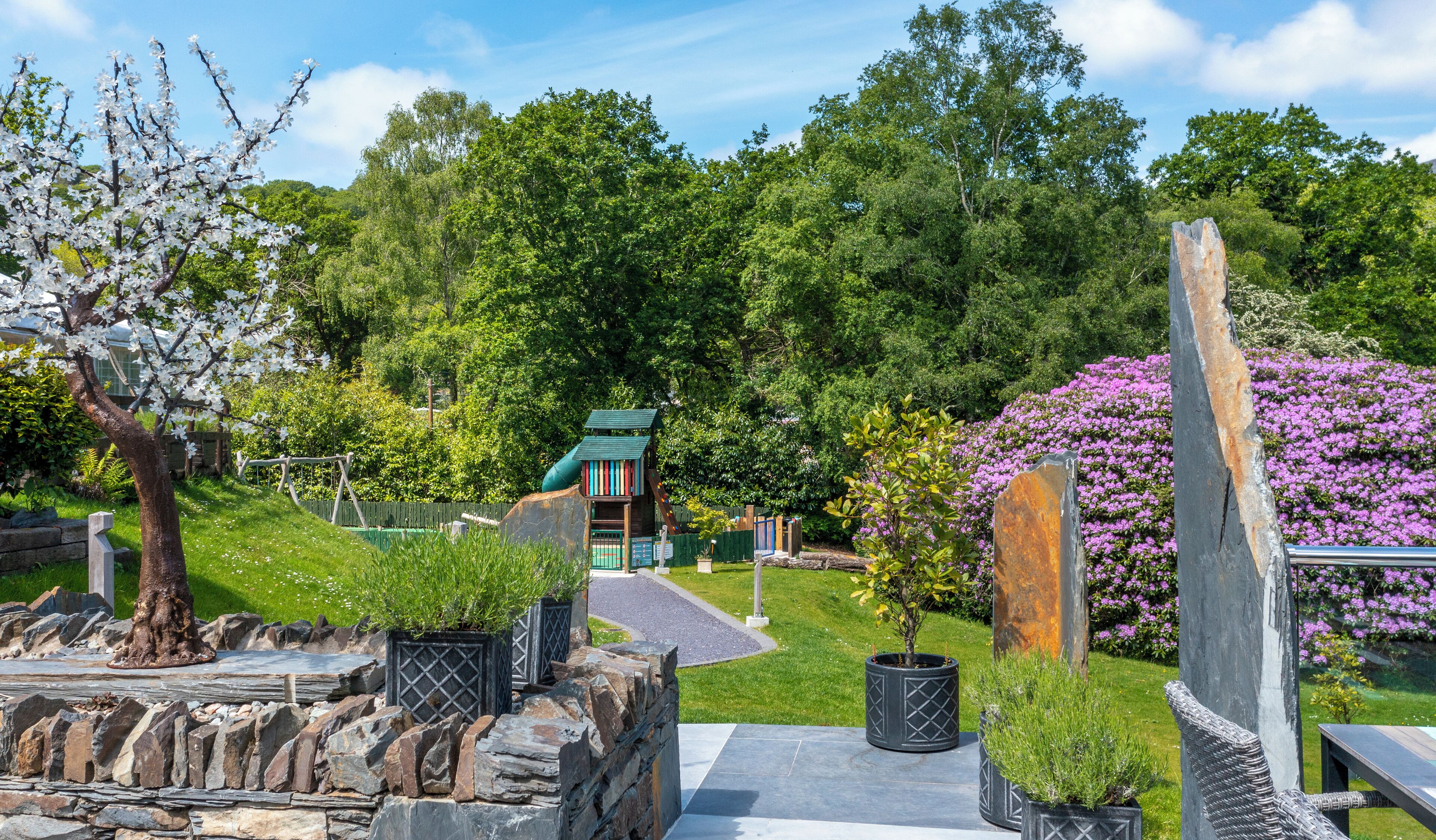 A garden with a stone wall, a wooden bench, and a small wooden structure with a green roof.
