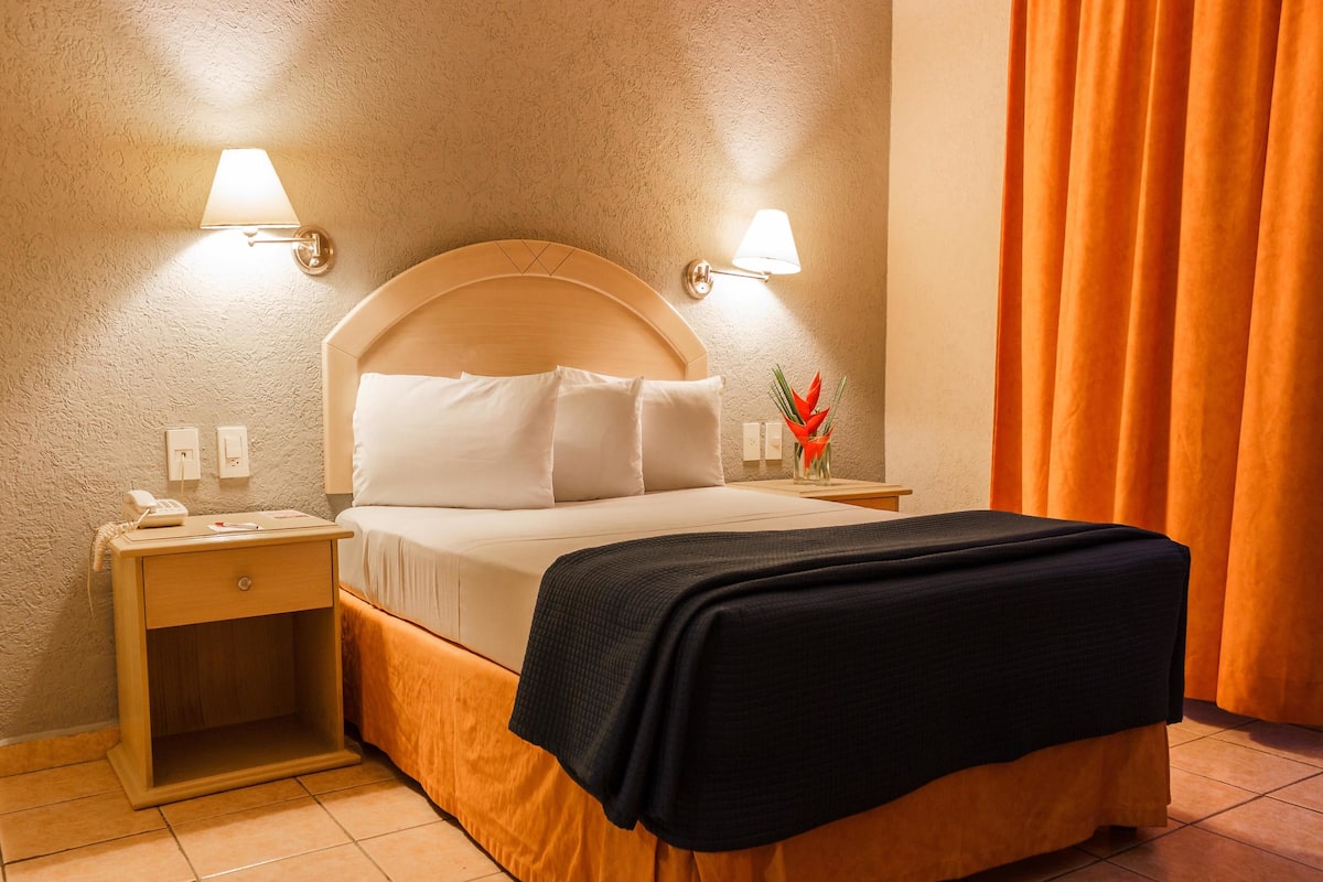 A hotel room with a bed, nightstand, lamp, and a vase with flowers.