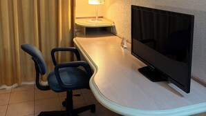 A hotel room with a television on a stand, a desk, a chair, and a lamp.