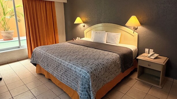A hotel room with a bed, two bedside lamps, a nightstand, a window with curtains, and a potted plant.