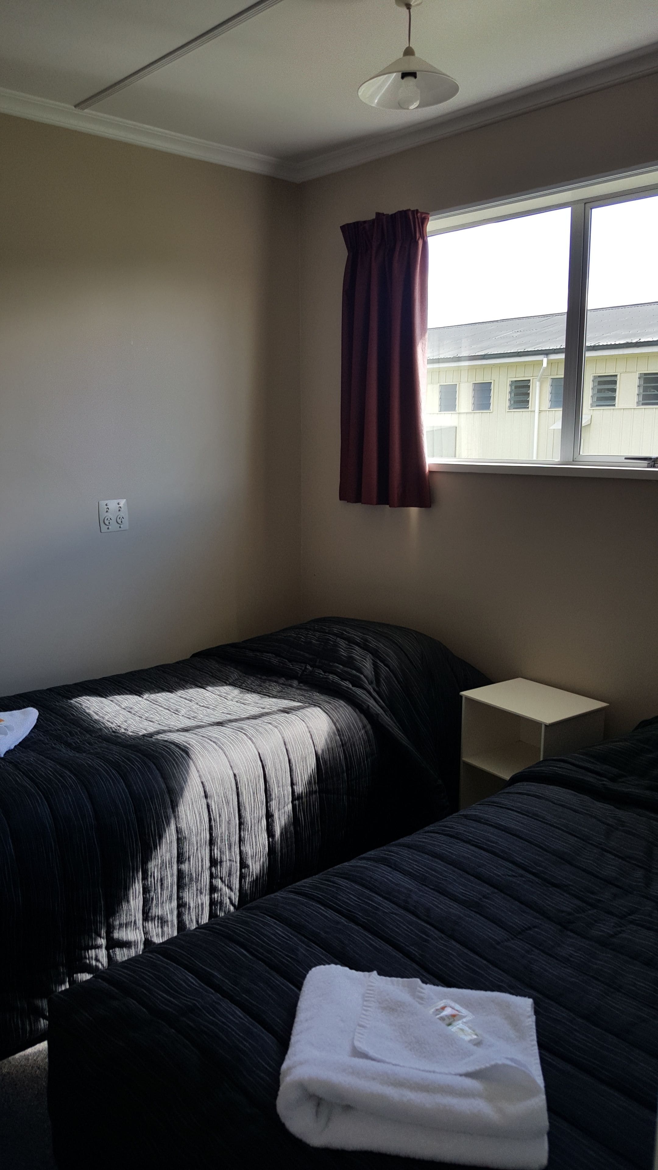 A room with two beds, a window with a curtain, a bedside table, and a ceiling light.