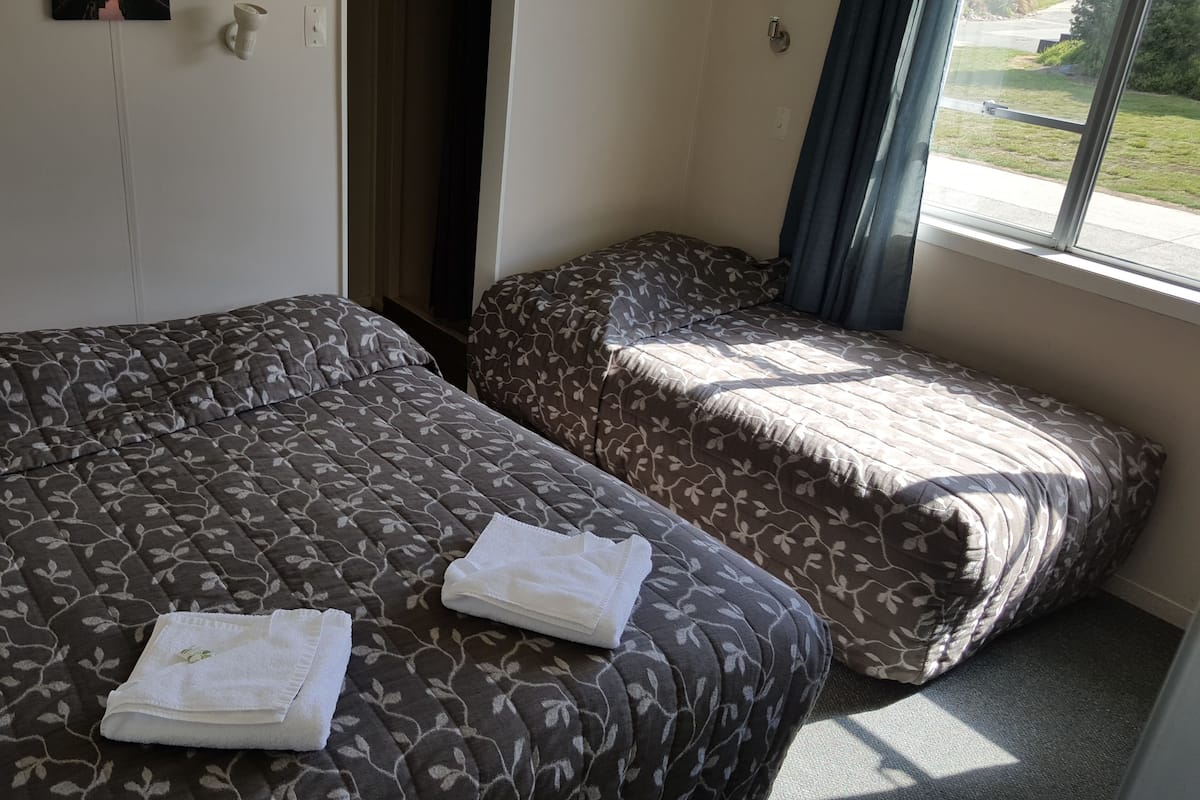 Ein Hotelzimmer mit zwei Betten, jeweils mit einem gemusterten Überwurf. Es gibt ein Fenster mit Vorhängen, eine Wandleuchte und eine Tür.