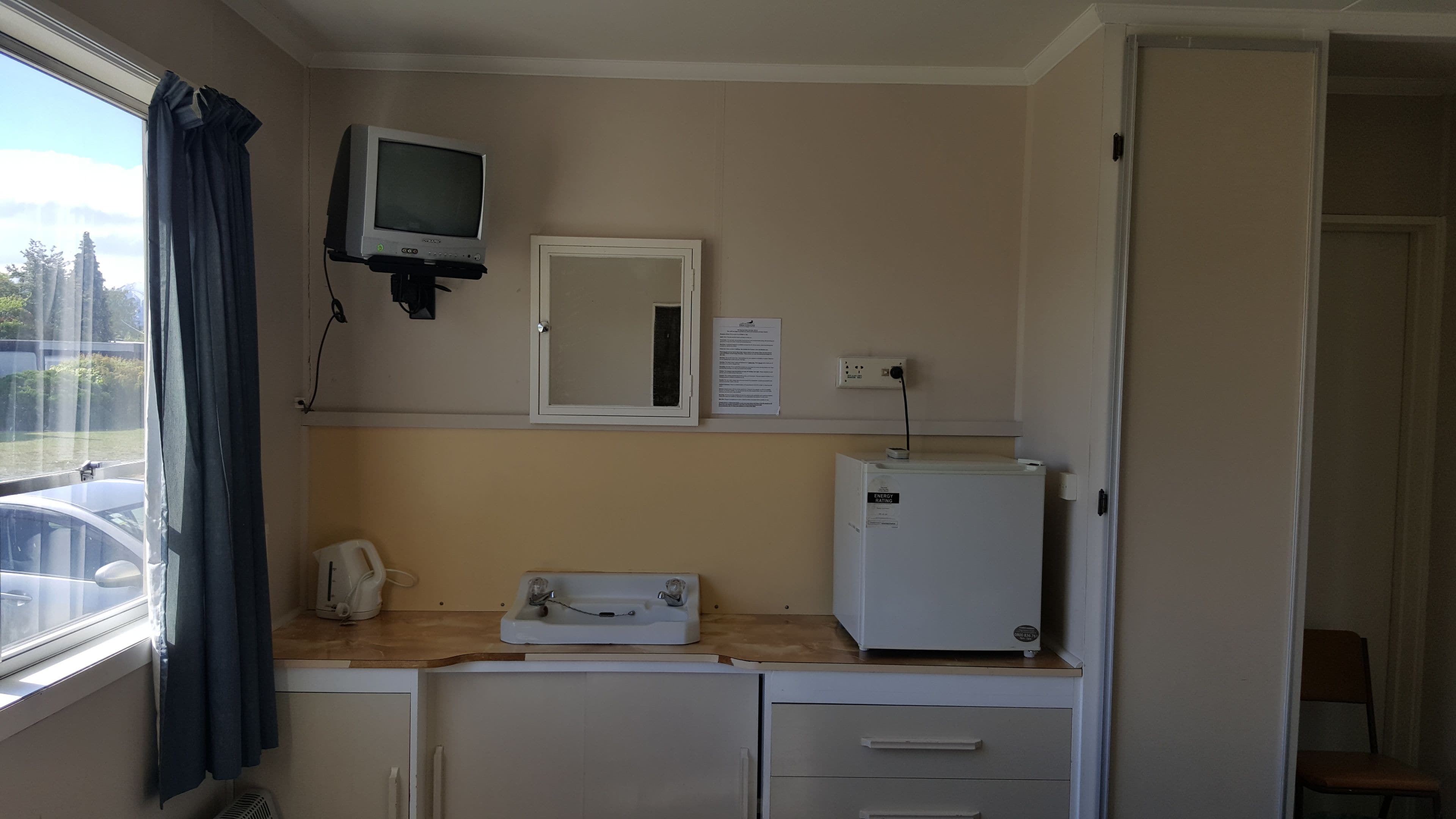A compact room with a kitchenette, a small refrigerator, a microwave, a telephone, a mirror, and a television mounted on the wall.