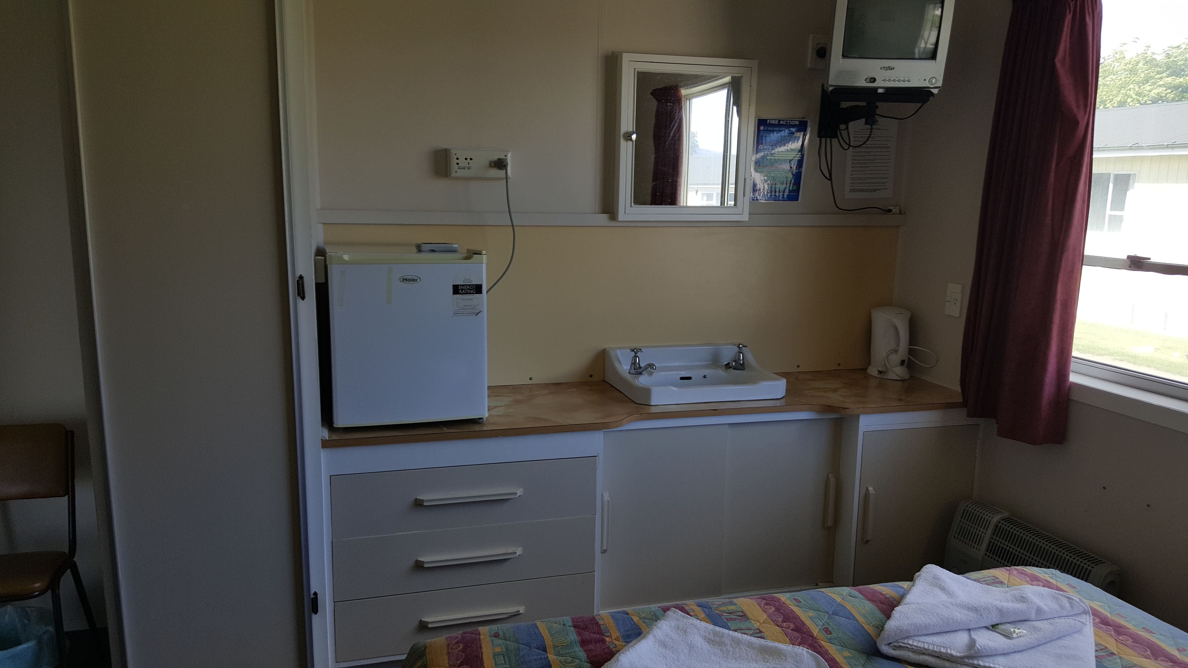 Quarto compacto com cama, mesa de cabeceira pequena, aquecedor embutido, janela com cortinas e televisão fixada na parede.