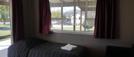 A room with a bed, a window with curtains, and a view of a parking lot and trees.