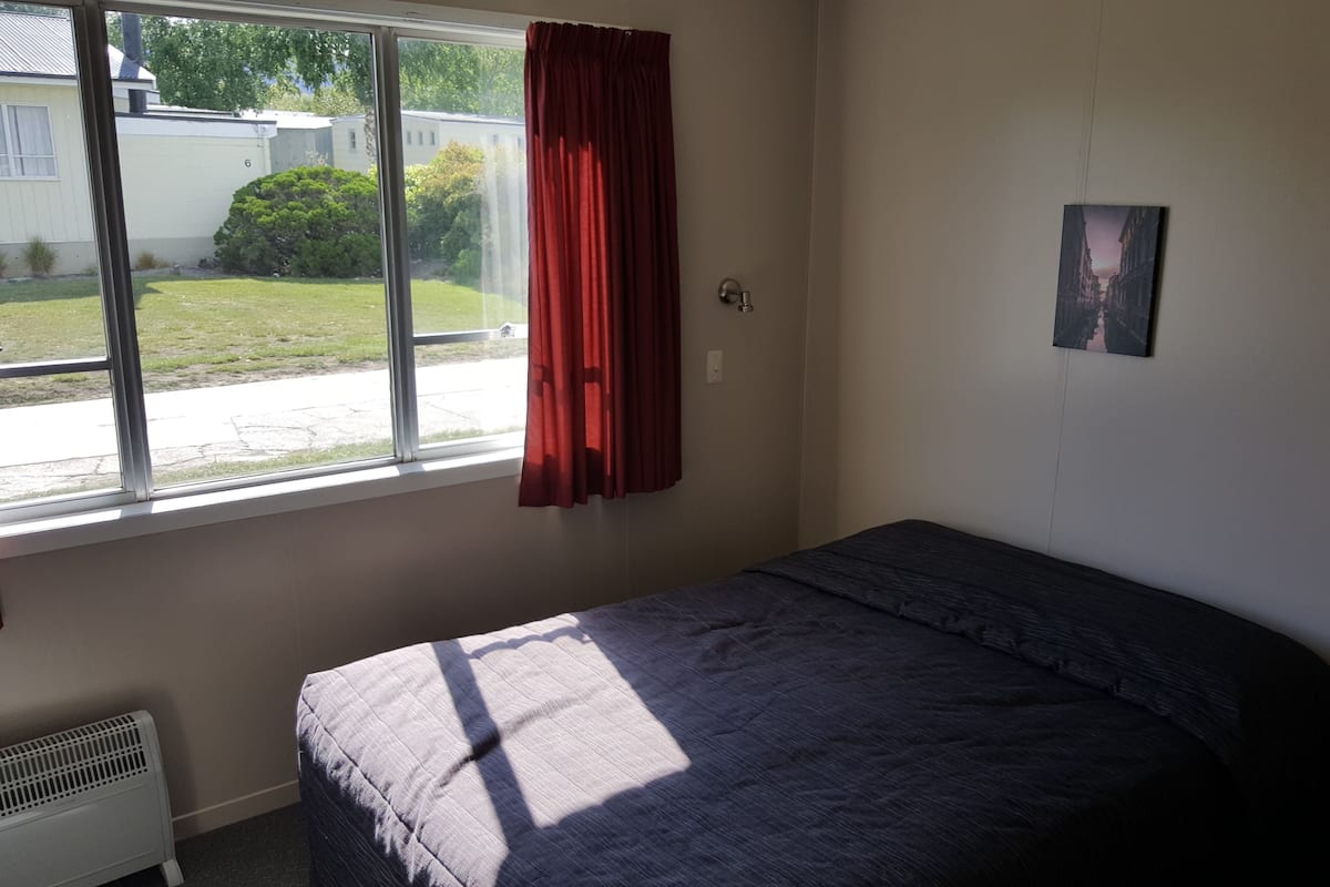 Ein Schlafzimmer mit einem Bett, einem Fenster mit rotem Vorhang, einem Heizkörper und einem an der Wand hängenden Bild.