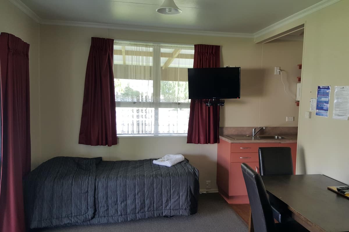 Ein kleines Zimmer mit Bett, einer kleinen Küchenzeile, einem Fernseher und einem Fenster mit Vorhängen.